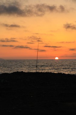 Güneş ufka doğru batarken, altın renkler gökyüzünü boyar. Kıyıda bir balıkçı oltası dik durur. Okyanus kıyısında sakin bir günün sonunu işaret eder..