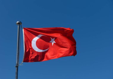 Canlı bir Türk bayrağı bulutsuz, masmavi bir gökyüzünün altındaki bayrak direğinin üzerinde zarif bir şekilde dalgalanıyor. Güneş kırmızı kumaşı aydınlatır, canlı rengini ve ikonik hilal ve yıldızını gösterir..