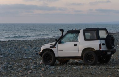 Gaziveren Kıbrıs Rum Kesimi 16.04.2025 engebeli bir SUV çakıl taşlarıyla döşenmiş bir plaja parketmiş ve hava kararırken pastel bir gökyüzünün altında dalgalar yavaşça kıyıya vuruyordu. Deniz kenarında keşif ve rahatlama için mükemmel bir an..