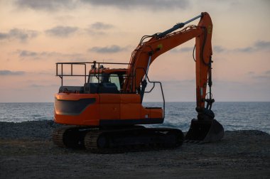 Suyun kenarında, canlı bir günbatımına karşı siluet oluşturan ağır makineler, yaklaşan kıyı inşaat görevlerine hazırlık niteliğinde..