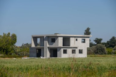 Yeşil bir alana yerleştirilmiş, minimalist beton yapı tamamlanmamış, mavi gökyüzü çarpıcı bir zemin sunuyor. Doğa, izolasyonunu vurguluyor..