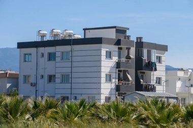 Çağdaş bir apartman yapısında, açık mavi gökyüzünün altında yemyeşil palmiye ağaçlarıyla çevrili renkli çamaşırların rüzgarda dalgalandığı balkonlar yer almaktadır..