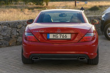 Gaziveren Kıbrıs Rum Kesimi 16.04.2025 Şık kırmızı bir spor araba kırsal taştan bir duvarın yanında duruyor, arka planda açık bir gökyüzünün altında gün batımında uzanan altın tarlalar var..