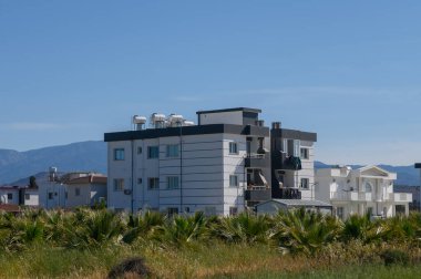 Açık mavi gökyüzü, canlı yeşilliklerin arasına yerleştirilmiş bir dizi modern yerleşim binasını ön plana çıkarıyor. Çevresindeki manzarada palmiye yaprakları rüzgarda hafifçe sallanıyor..