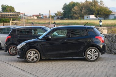 Gaziveren Kıbrıs Rum Kesimi -16.04.2025. Parlak siyah bir sedan, yemyeşil tarlalarla çevrili diğer iki aracın yanına park etmiş ve güneş batarken huzurlu bir ortam yaratmış..