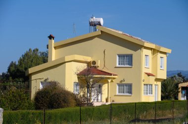 Geniş pencereleri ve eğimli çatısı olan büyüleyici sarı bir ev, yemyeşil çimenler ve ağaçlarla çevrili, huzurlu bir çevrede duruyor. Parlak güneş ışığı davetkar görünüşünü arttırır..