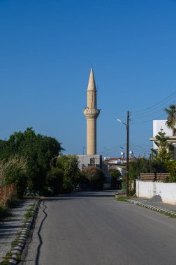 Huzurlu bir yolun üzerinde görkemli bir şekilde ince minare kuleler, yemyeşil ve konut evleriyle dolu. Açık mavi gökyüzü, sahnenin sakin atmosferine ekleniyor..