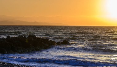 Canlı bir günbatımı, gölün durgun yüzeyini yansıtan, dağların üzerine sıcak renkler saçar. Siluetli ağaçlar sahneyi çevreleyerek, akşamın huzurlu atmosferini güçlendiriyor..