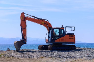 Akdeniz 'in mavi suları ve engebeli kıyı şeridi tarafından çerçevelenen Kıbrıs' taki bir kıyı şeridinde turuncu bir kazıcı boşta duruyor