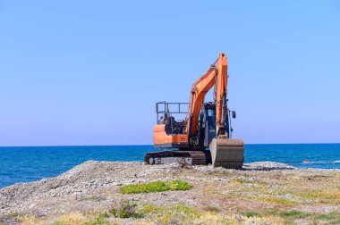 Kıbrıs 'ta Akdeniz yakınlarında faaliyet gösteren turuncu kazıcı, mavi gökyüzü ve deniz zeminine karşı kıyı gelişimi ile meşgul.