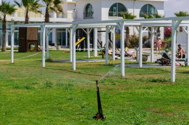 Sıcak ve güneşli bir günde, Kıbrıs 'taki yeşil çimlerin üzerine serinletici su püskürtülmesi. Akdeniz bahçe hayatı, doğal güzellik ve sürdürülebilir manzara bakımı.