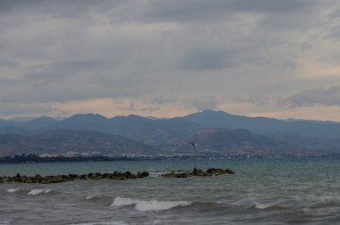 Kıbrıs kıyı şeridinde gün batımında, arka planda dağlarla birlikte, sakin bir sahilden görüldü. Sıcak renkler, deniz dalgaları ve doğal güzellik sakin bir sahil manzarası yaratır..