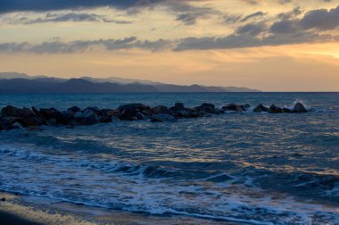 Sıcak altın güneş ışığı, gün batımında Kıbrıs 'ın kayalık kıyılarını aydınlatıyor. Sakin dalgalar, parlayan gökyüzü ve deniz kenarındaki sakin ada atmosferi..