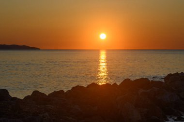 Güneş ufukta batarken, altın ve turuncu renkler sakin denizi aydınlatarak huzurlu bir ortam yaratıyor. Siluetli kayalar bu güzel anı çerçeveler, sükuneti davet eder..