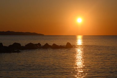 Güneş, dingin okyanusun üzerine sıcak turuncu ve sarı renkler saçarak yavaş yavaş alçalıyordu. Dalgalar yavaşça karanlık kayalara çarpar. Alacakaranlık yaklaşırken huzurlu bir ortam yaratır..