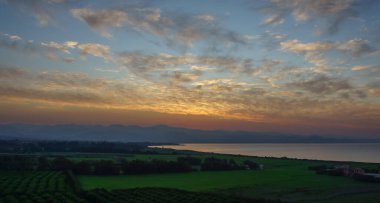 Gün ışığı sönerken, yeşil alanlar ve gölgeli dağlarla çevriliyken huzurlu bir göl turuncu ve mavi renkli canlı renkleri yansıtır. Doğa sakinliği sahneyi büyülüyor..