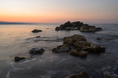 Güneş ufukta batarken yumuşak dalgalar engebeli kayalara çarpar, gökyüzünü turuncu ve mor renkler içinde huzurlu bir sahile boyar, sükuneti ve tefekkürü davet eder..