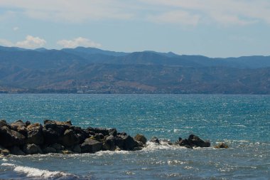 Sakin sular gün ortası güneşini yansıtır. Yumuşak dalgalar engebeli kayalara çarpar. Uzakta, dağlar görkemli bir şekilde yükselirler, sakin bir sahil atmosferi yaratarak rahatlamayı davet ederler..