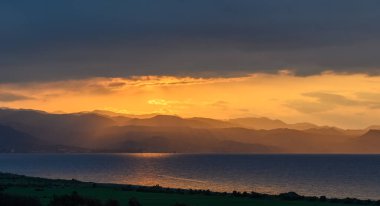 Güneş görkemli dağların ardında batarken altın renkler sakin okyanustan yansıyor. Dramatik bulutlar gökyüzüne derinlik katar, alacakaranlığın huzurlu güzelliğini arttırır..