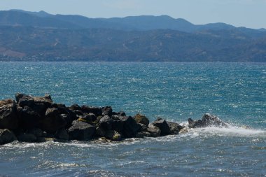 Sakin sular, uzaktaki sisli dağlarla buluşmak için uzanıyor. Dalgalar, deniz kenarındaki huzurlu bir günde güneş ışığını yansıtan yıpranmış kayalara hafifçe çarpar..