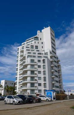 Çarpıcı beyaz bir gökdelen canlı bir şehir ortamının ortasında dimdik ayakta duruyor. Aşağıda, park halindeki arabalar ön planda sıralanırken, gökyüzü şehir için dinamik bir ortam yaratıyor..