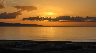 Güneş barışçıl bir okyanusun üzerinde batarken canlı renkler gökyüzünü boyuyor, suya altın yansımalar yapıyor ve dingin bir atmosfer yaratıyor. Uzak tepelerin siluetleri güzelliği arttırır.