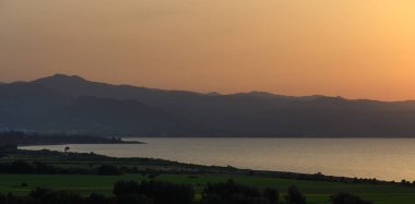 Gün ışığı söndükçe, sakin sular günbatımının turuncu ve altın renklerini yansıtıyor, siluetli dağlar ve canlı yeşil alanlar tarafından çerçevelenmiş, huzurlu bir akşam atmosferi yaratıyor..