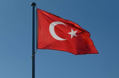 Rüzgarda dalgalanan çarpıcı bir Türk bayrağı, beyaz hilal ve yıldızla zıtlaşan koyu kırmızı rengini sergiliyor. Açık mavi gökyüzü ulusal gururu kutlamak için mükemmel bir zemin oluşturur..