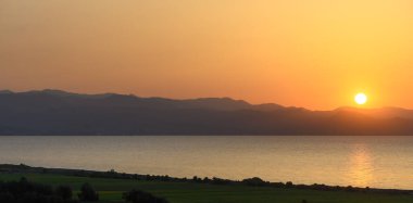 Gündüz geceye geçerken, parlak bir günbatımı dingin suların üzerine sıcak altın bir parıltı saçar. Uzak dağlar ufku çerçeveler, bu doğal ortamda barışçıl bir atmosfer yaratır..
