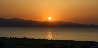 Güneş batarken, gökyüzü portakal ve altının sıcak gölgelerine dönüşür, sakin suların ve siluetli dağların üzerine huzur dolu bir parıltı yayar, nefes kesici bir akşam manzarası yaratır..