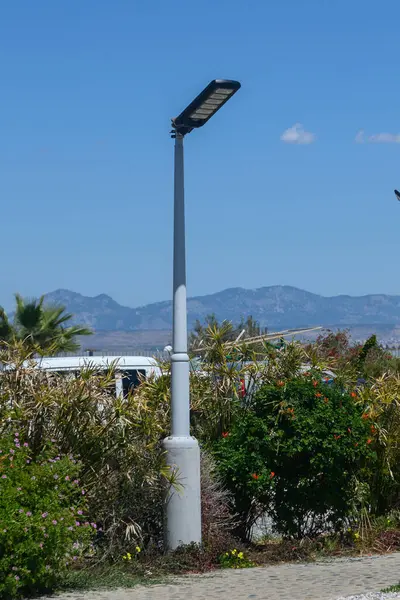 Çağdaş bir sokak lambası yemyeşil çimlerin üzerinde yükselir, parlak bir güneşin altında ışık saçar. Görkemli dağlar, bölgenin doğal güzelliğini artırmak için çarpıcı bir zemin oluşturur..