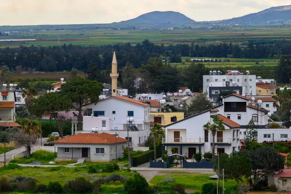 Kıbrıs 'taki huzurlu bir köy, bulutlu bir gökyüzü altında yemyeşil tarlalar ve dağlarla çevrili beyaz yıkanmış evler ve tanınmış bir minareyi gözler önüne seriyor ve yerel cazibe ve sükuneti yakalıyor..