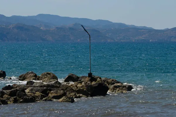 Dalgalar, güneşin ısıttığı kayaları hafifçe okşar. İnce bir direk, durgun bir kıyı bölgesini işaret ederken. Arka planda dağlar görkemli bir şekilde yükselir, barışçıl bir atmosfer yaratır..