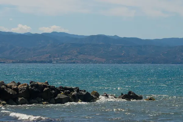 Sakin sular gün ortası güneşini yansıtır. Yumuşak dalgalar engebeli kayalara çarpar. Uzakta, dağlar görkemli bir şekilde yükselirler, sakin bir sahil atmosferi yaratarak rahatlamayı davet ederler..