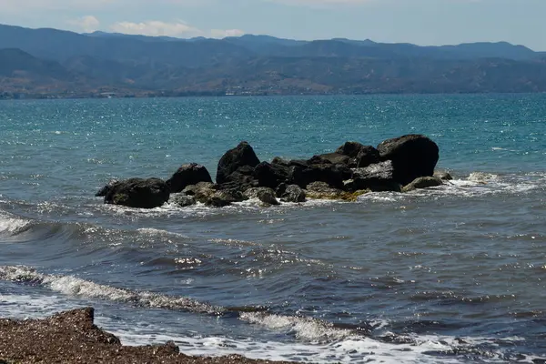 Dalgalar açık okyanus kıyısındaki karanlık kayalara nazikçe dokunur. Güneşli bir gökyüzünün altında uzak tepeler ve dağlar doğayla huzurlu bir bağ oluşturur..