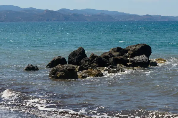 Sakin okyanus dalgaları engebeli taşlara nazikçe sarılarak sakin bir atmosfer yaratıyor. Arka planda, uzak dağlar parlak mavi bir gökyüzünün altında dikilir ve manzarayı güzelleştirir..