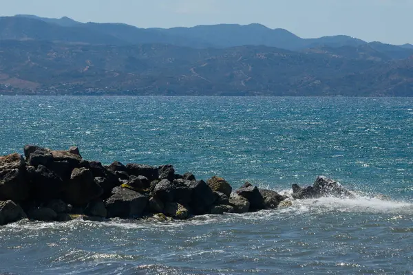 Sakin sular, uzaktaki sisli dağlarla buluşmak için uzanıyor. Dalgalar, deniz kenarındaki huzurlu bir günde güneş ışığını yansıtan yıpranmış kayalara hafifçe çarpar..