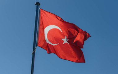 İkonik hilal ve yıldızıyla süslenmiş parlak bir Türk bayrağı, ulusal gurur ve birliği simgeleyen canlı mavi gökyüzünün altında enerjik bir şekilde dalgalanır..