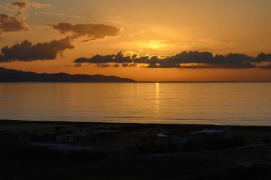 Altın güneş ışığı sakin suları yansıtıyor. Güneş uzak tepelerin ardından inerken sakin kıyı şeridi boyunca huzurlu bir alacakaranlık atmosferi yaratıyor. Doğanın güzelliği huzur verir..