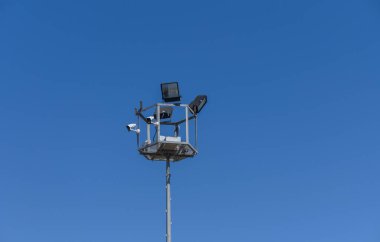 Kameralar ve projektörlerle donatılmış uzun bir gözetleme kulesi parlak mavi gökyüzünün önünde duruyor. Çağdaş kurulumda güvenlik ve gözlem amaçlı modern teknoloji sergileniyor.