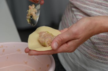 Sıcak bir mutfakta, yetenekli eller kaşıkla ince bir hamur çemberine doluyor, geleneksel hamur köftelerini şekillendiriyor. Atmosfer yaratıcılık ve aşçılık tutkusu yayar..