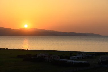 Güneş sakin suların altına batarken turuncu ve sarı renkli canlı renkler gökyüzünü dolduruyor. Uzak dağlar sakin bir zemin oluşturur, yemyeşil evlerle çevrili evler, huzuru çağrıştırır..