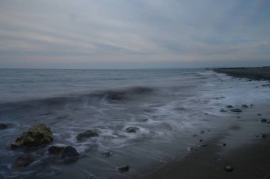 Yumuşak dalgalar çakıllı bir kumsalda yuvarlanırken, alacakaranlık gökyüzü su üzerinde sakin bir hava yaratırken huzurlu bir ritim yaratıyor. Ufuk, karanlık gökyüzü ile kusursuz bir şekilde harmanlanır..