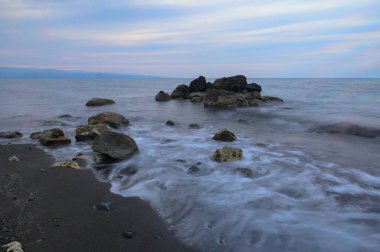 Alacakaranlık yaklaşırken dalgalar sakin bir sahil boyunca kayaların üzerinde usulca dalgalanır. Akşam gökyüzü yumuşak bir pastel palet tutuyor, alacakaranlıkta rahatlatıcı bir atmosfer yaratıyor..
