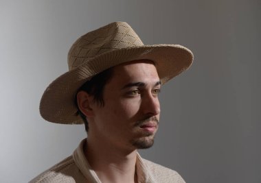 Genç bir adam şık bir hasır şapka giyerken düşünceli bir şekilde bakıyor. Yumuşak ışık, yüz hatlarını ön plana çıkarıyor. Minimalist bir ortamda iç gözlem atmosferi yaratıyor..