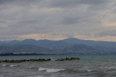 Kıbrıs kıyı şeridinde gün batımında, arka planda dağlarla birlikte, sakin bir sahilden görüldü. Sıcak renkler, deniz dalgaları ve doğal güzellik sakin bir sahil manzarası yaratır..