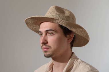 Hafif hasır şapka giyen bir adam sessizce oturur, düşüncelere dalmış. Yumuşak nötr renklerle çevrili, sakin atmosferi arttıran içsel bir ifadesi var..