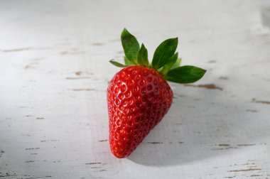 Tek bir olgun çilek eskimiş beyaz ahşap bir yüzeye dayanır, canlı kırmızı tonu yeşil yapraklarla mükemmel zıtlaşır. Doğal güzellik tazeliğini vurgular..
