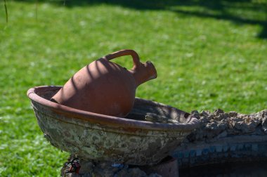 Güzel işlenmiş bir kil tenceresi suyla dolu bir taş havzanın kenarına nazikçe dayanıyor. Bereketli yeşillik sahneyi sararken güneş ışığı yumuşak gölgeler bırakır ve sakin bir atmosfer yaratır..