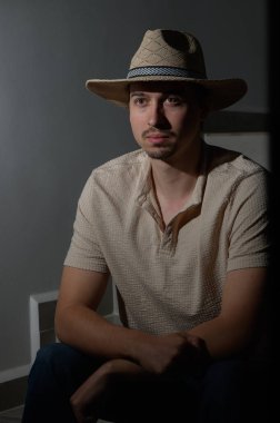 Hasır şapkalı genç bir adam, minimalist bir ortamda sessizce oturur. Yüzündeki ılık ışık, onun özelliklerini nazikçe vurgular ve yansımasını gösteren samimi bir atmosfer yaratır..